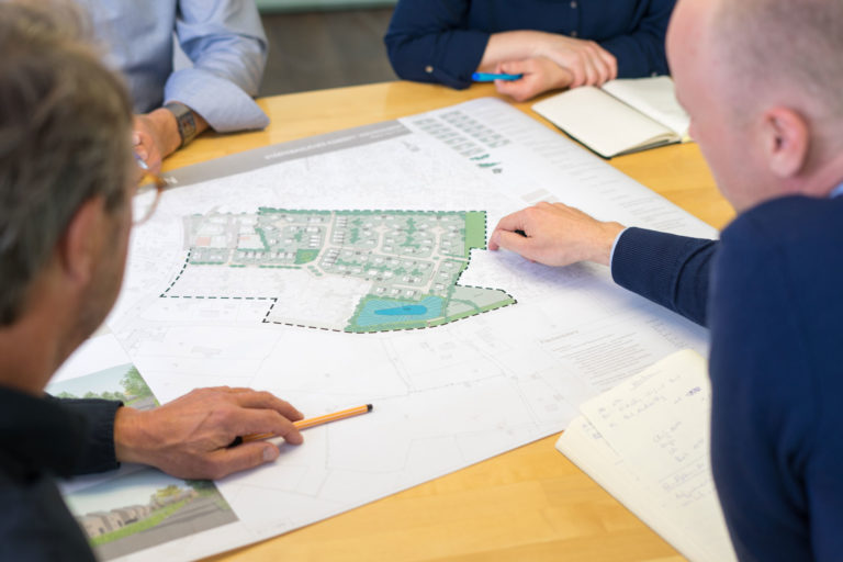 Mehrere Personen sitzen an einem Tisch und beraten über einen ausgebreiteten Lageplan einer Wohnsiedlung mit markierten Grundstücksgrenzen, Grünflächen und einem kleinen Teich; Hände zeigen auf Details, ein Bleistift und Notizbücher liegen daneben.