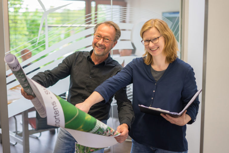 Zwei lächelnde Personen — ein Mann und eine Frau — stehen in einem Büro vor einer Glaswand mit weißem Streifenmuster und rollen gemeinsam große gedruckte Plakate aus, während die Frau ein Klemmbrett hält.