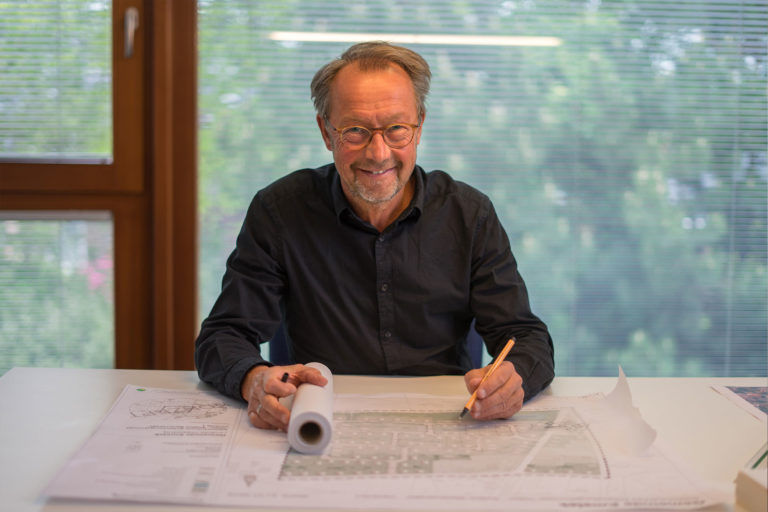 Älterer Mann mit Brille und schwarzem Hemd sitzt lächelnd an einem Tisch, hält eine aufgerollte Bauzeichnung und zeichnet mit einem Bleistift auf ausgebreiteten Plänen vor einem Fenster mit Jalousien und grünem Außenblick.