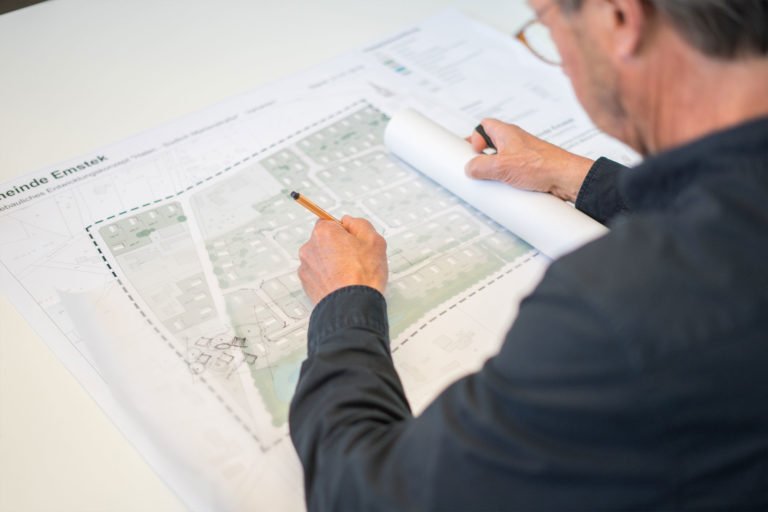 Person von hinten über einem ausgebreiteten Bebauungsplan, zeichnet mit Bleistift auf einer durchsichtigen Folie und hält eine aufgerollte Planseite; auf dem Plan sind ‚Gemeinde Emstek‘, Grundstücksgrenzen und Grünflächen erkennbar.