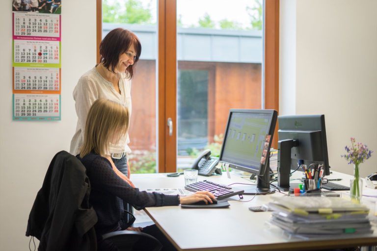 Zwei Frauen in einem hellen Büro an einem Schreibtisch mit zwei Monitoren, eine sitzt und bedient die Maus, die andere steht lächelnd daneben; im Hintergrund große Fenster, ein Wandkalender und auf dem Tisch Stapel Unterlagen, Stifte und eine kleine Blumenvase.
