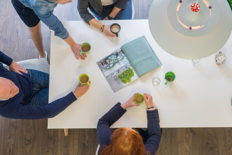 Vogelperspektive auf vier Personen um einen weißen Tisch, die jeweils eine grüne Tasse Kaffee halten; in der Mitte liegt eine aufgeschlagene Zeitschrift, rechts oben hängt eine große Hängeleuchte und steht eine Schale mit Zuckerwürfeln, der Raum hat dunklen Holzboden.