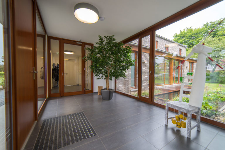 Heller Eingangsbereich mit großen Holzrahmen-Fenstern und Glasfront, grauem Fliesenboden und Fußmatte, einer großen Topfpflanze (Ficus) in der Mitte sowie einem kleinen weißen Windrad auf einem Hocker; durch die Fenster Blick auf einen Backsteinbau und Garten.