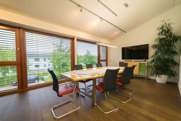 Heller Besprechungsraum mit großem rechteckigem Holztisch, sechs Stühlen mit schwarzen Rücken und roten Sitzen, verstreuten Papieren auf dem Tisch, großen Fensterfronten mit horizontalen Lamellen, hängender Deckenleuchte, Wandfernseher auf einem Sideboard und hoher Topfpflanze in der Ecke.