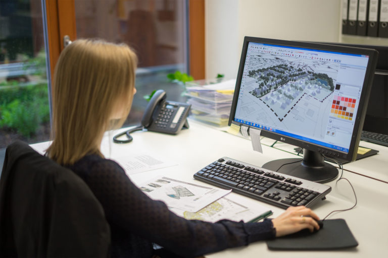 Person sitzt an einem Büroarbeitsplatz und betrachtet auf dem Monitor ein 3D-Stadtmodell mit Häusern und Bäumen; auf dem Schreibtisch liegen Pläne, Tastatur, Maus und ein Telefon.