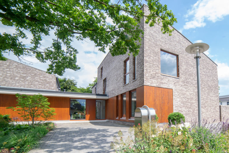 Modernes zweistöckiges Gebäude mit hellen Ziegeln und vertikalen Holzverkleidungen, großen Fenstern, gepflastertem Vorplatz und Blumenbeeten, davor ein Laternenmast und überhängende Baumzweige an einem sonnigen Tag.