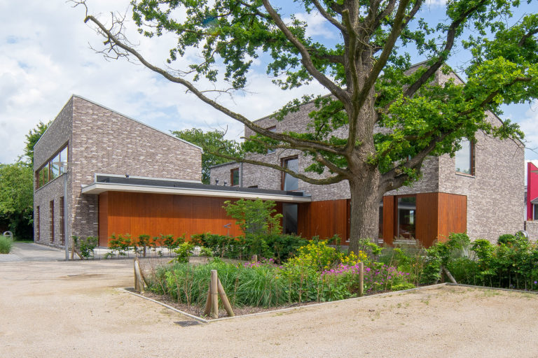 Modernes, kubisches Haus aus hellgrauen Ziegeln mit dunklen Holzverkleidungen, davor ein großer Eichenbaum, ein bepflanztes Blumenbeet und ein Kiesplatz.