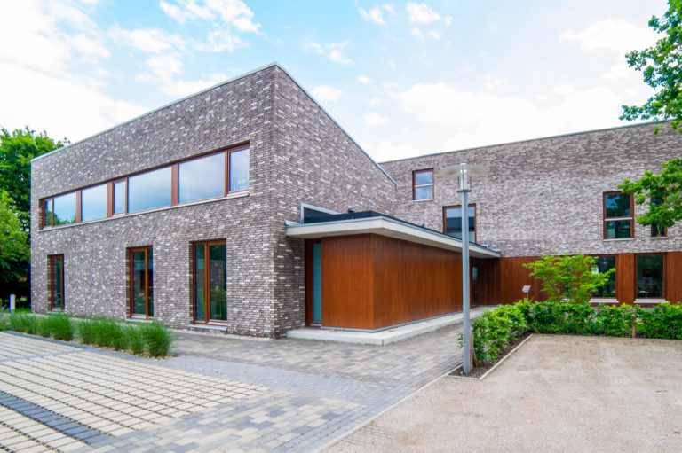 Eckansicht eines modernen, kubischen zweistöckigen Gebäudes mit hellgrauen Ziegeln und Holzverkleidung, großen rechteckigen Fenstern, überdachtem Eingang, gepflastertem Vorplatz, Laterne und niedrigen Sträuchern unter blauem Himmel.