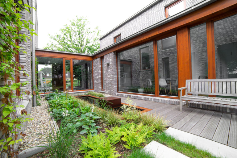 Innenhof eines modernen Einfamilienhauses mit grauer Klinkerfassade und großformatigen Holzrahmen-Fenstern, einem Holzdeck mit Parkbank neben einem rechteckigen Teich sowie Beet mit Farnen, Stauden und Kiesweg.