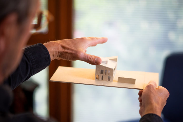 Hände einer Person halten ein kleines Karton-Architekturmodell eines modernen Hauses mit schrägem Dach und rechteckigen Fenstern auf einer flachen Holzplatte vor unscharfem Innenraumhintergrund.