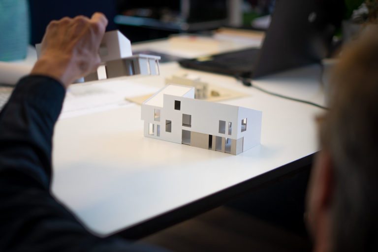 Maßstabsmodell eines modernen, zweigeschossigen weißen Hauses mit rechteckigen Fensteröffnungen auf einem weißen Tisch, im Hintergrund eine Hand, die ein weiteres Modell hält, sowie unscharfe Arbeitsmaterialien und ein Laptop.