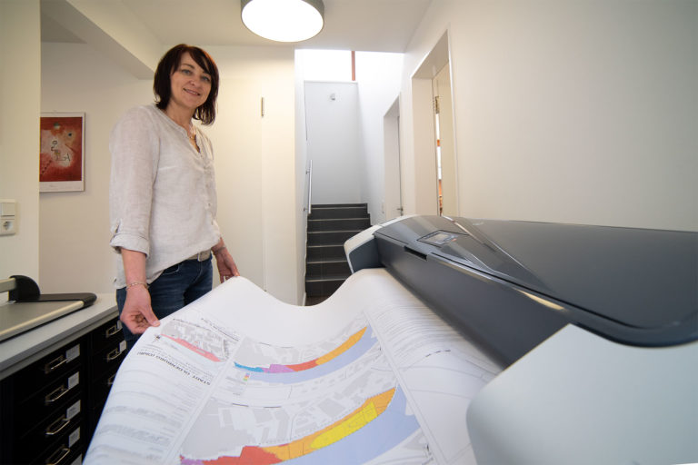 Eine lächelnde Frau in heller Bluse steht in einem hellen Büroflur neben einem großformatigen Drucker und zieht einen großen, farbig kodierten Stadtplan/Grundriss aus dem Gerät.