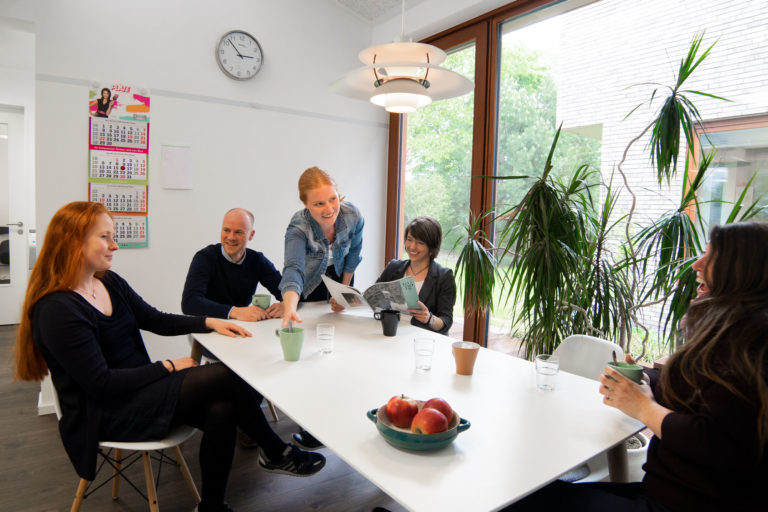 Fünf Personen in einem hellen Besprechungsraum an einem weißen Tisch; eine Frau lehnt sich lächelnd vor und reicht eine Tasse, die anderen sitzen mit Bechern, auf dem Tisch eine Schale mit Äpfeln und im Hintergrund große Fenster mit Grünpflanzen sowie eine Uhr und ein Kalender an der Wand.