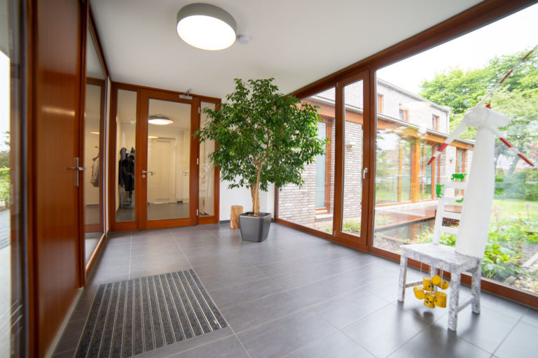 Heller Eingangsbereich mit großen Holzrahmen-Glasfenstern und -türen, grauem Fliesenboden, einem großen Topfbaum in der Mitte, einer Fußmatte links und einem kleinen weißen Windrad auf einem Hocker rechts, mit Blick auf Garten und angrenzendes Backsteingebäude.