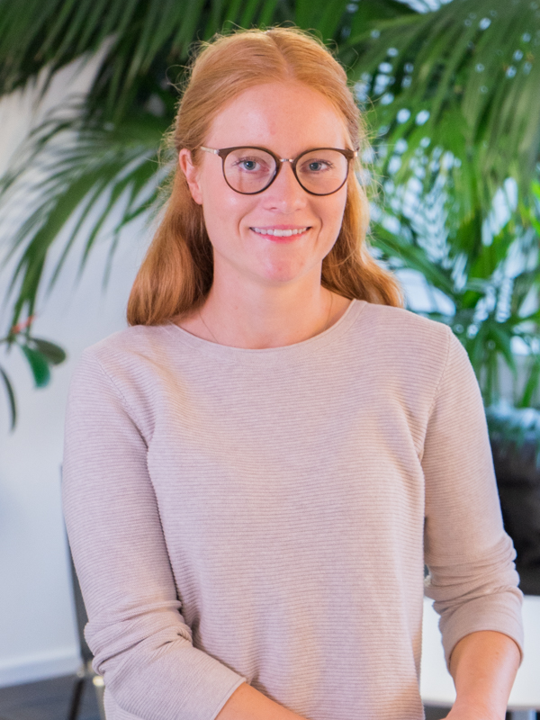 Frau mit roten Haaren und runder Brille, lächelnd in einem hellen, fein gerippten Pullover vor großen grünen Zimmerpflanzen.