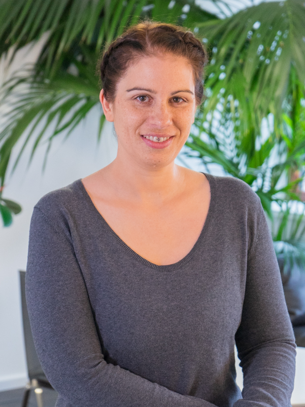 Lächelnde Frau mit geflochtenem Haar und grauem Pullover vor großen grünen Zimmerpflanzen in hellem Innenraum.