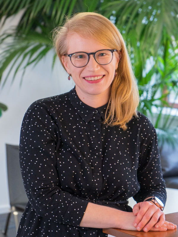 Lächelnde Frau mit Brille und schulterlangem blondem Haar, trägt ein schwarz-weiß gepunktetes Hemd und sitzt mit den Händen auf einem Tisch vor großen grünen Zimmerpflanzen.