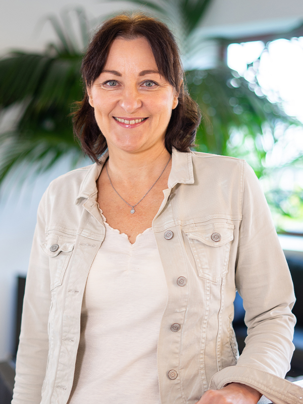 Lächelnde Frau mittleren Alters mit dunklen schulterlangen Haaren, in einer hellen Jeansjacke, weißem Top und einer kleinen silbernen Kette vor unscharfem Innenraum mit grünen Pflanzen.