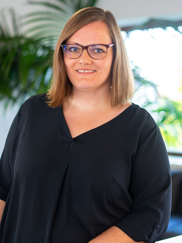 Porträt einer lächelnden Frau mit schulterlangem hellbraunem Haar und dunkler Brille, in einer schwarzen Bluse vor einem unscharfen Bürohintergrund mit Zimmerpflanzen und Fenster.