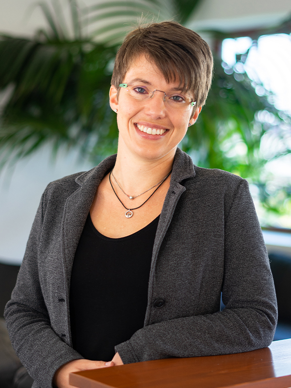 Lächelnde Person mit kurzen braunen Haaren und heller Brille, bekleidet mit grauem Blazer und schwarzem Top, lehnt mit den Händen auf einer Holzfläche vor einem unscharfen Innenraum mit großen grünen Pflanzen.