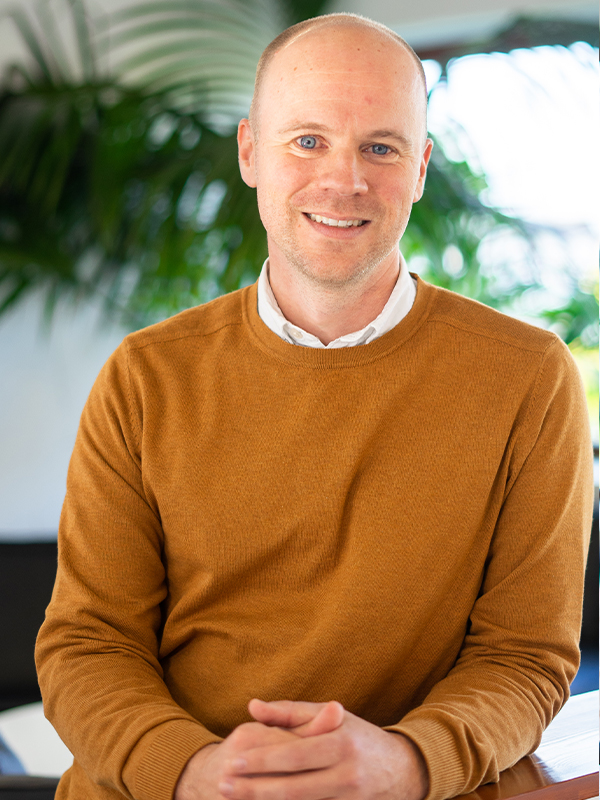 Porträt einer lächelnden, glatzköpfigen Person in einem senfgelben Pullover und weißem Hemd, die mit verschränkten Händen vor einer hellen, pflanzenverzierten Innenraumkulisse sitzt.