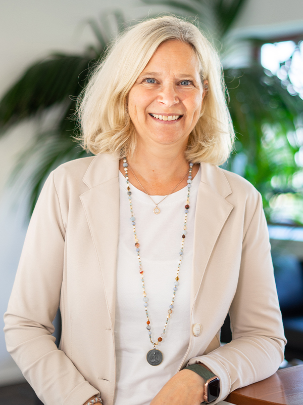 Lächelnde mittelalte Frau mit schulterlangem blondem Haar, die einen hellen Blazer, weißes Shirt, eine lange Perlenkette und eine Smartwatch trägt und vor einem unscharfen Hintergrund mit grünen Zimmerpflanzen steht.