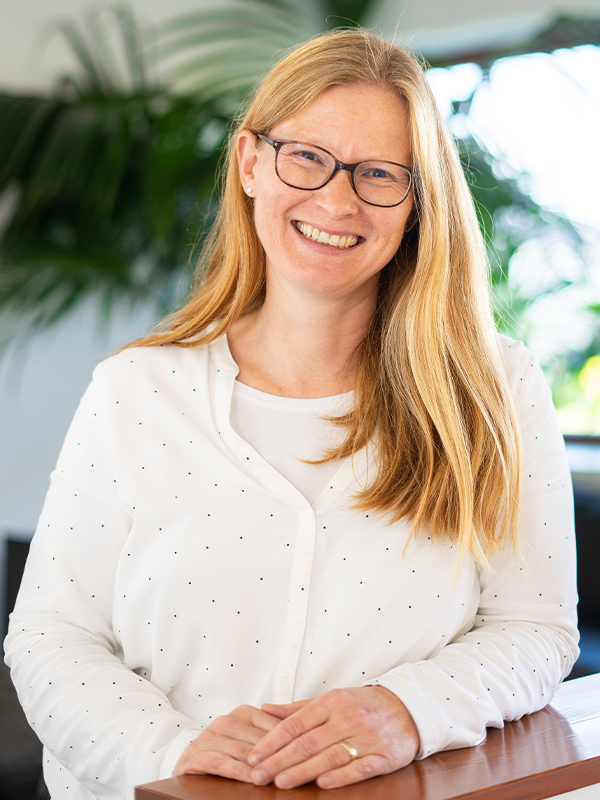 Lächelnde Frau mit langen blonden Haaren und Brille in einer weißen, leicht gepunkteten Bluse, die mit den Händen auf einer Holzoberfläche vor grünen Zimmerpflanzen posiert.