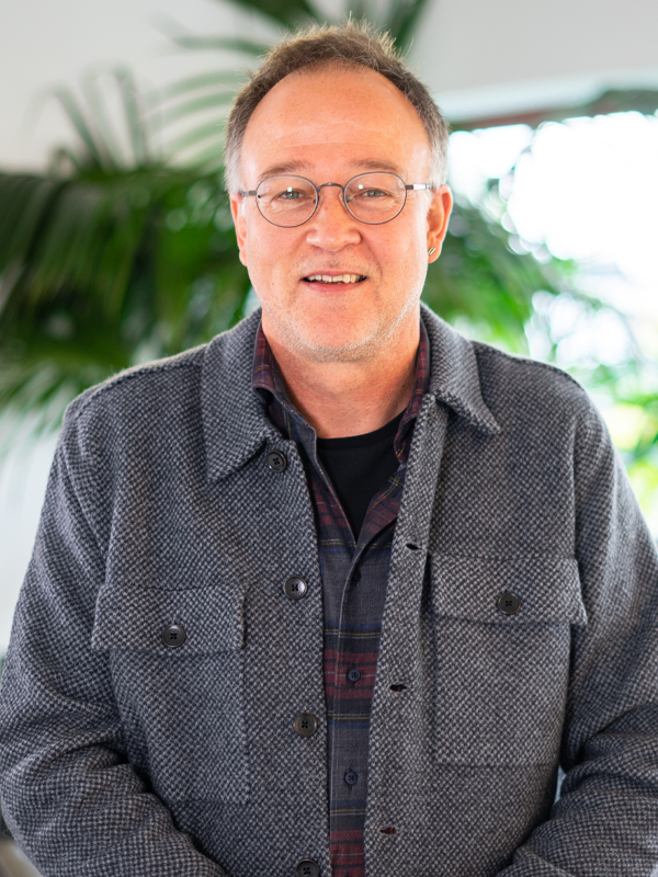 Lächelnder Mann mit runden Brillengläsern, kurzen leicht ergrauten Haaren und leichtem Bartstoppel, trägt eine graue, strukturierte Jacke über einem karierten Hemd und steht vor verschwommenen Grünpflanzen im Hintergrund.